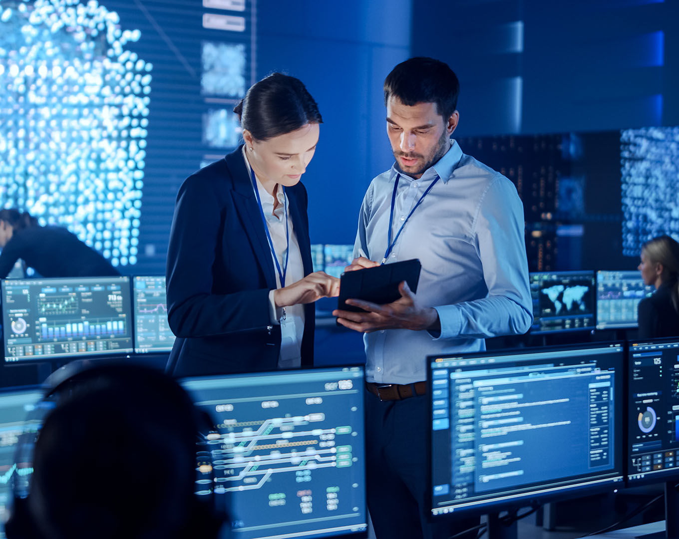 The image depicts two professionals, a man and a woman, standing in a high-tech control room or data centre. Both are dressed in business attire, wearing ID badges on lanyards, and appear to be collaborating while looking at a tablet. The background is filled with large digital screens displaying complex data, charts, and maps, suggesting a highly analytical or technical environment. The lighting is cool-toned, with a futuristic feel, indicating they are likely involved in cybersecurity, data analysis, or project management. The atmosphere conveys focus and teamwork in a cutting-edge professional setting.