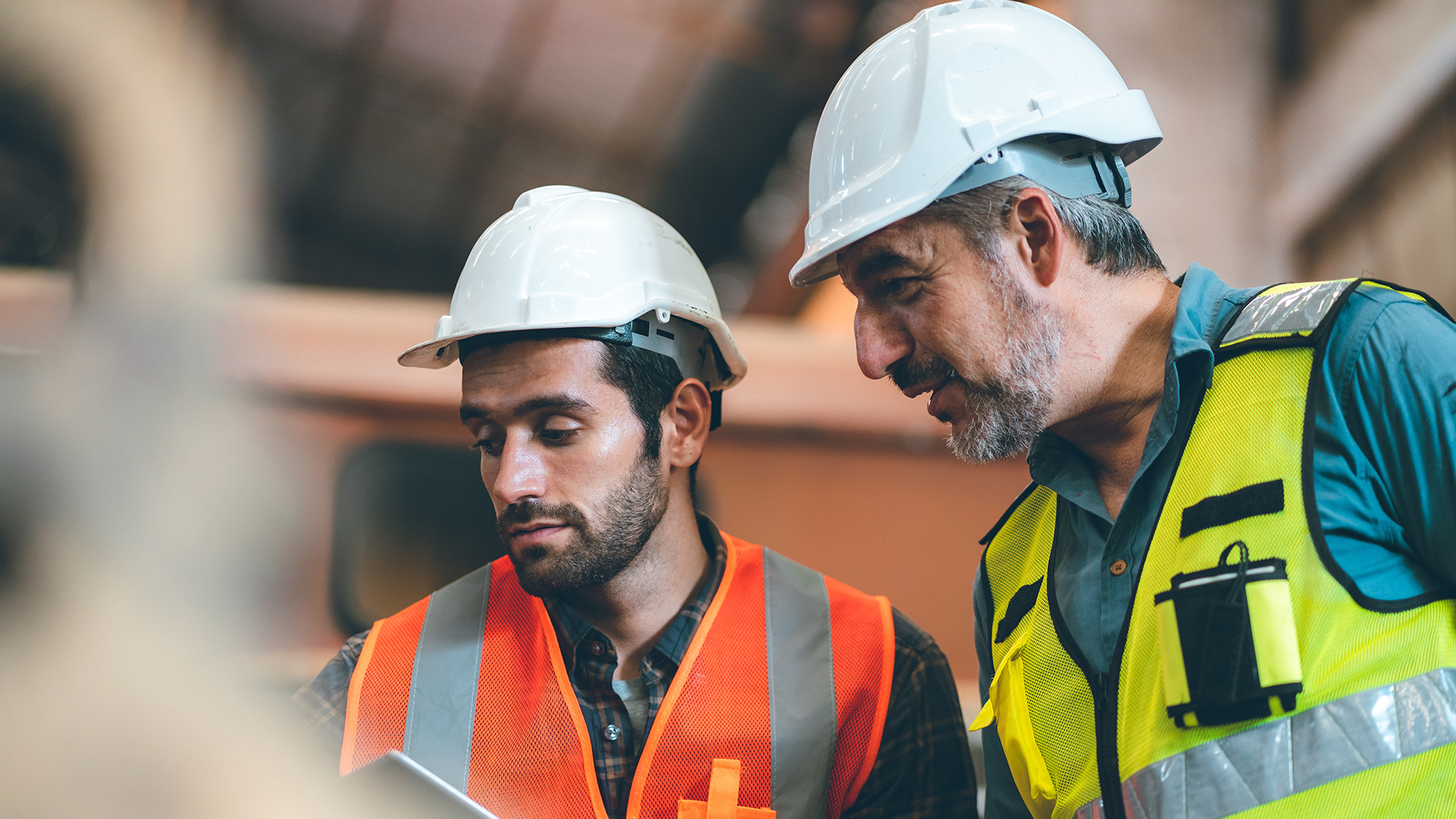 Two construction workers wearing hard hats and safety vests are having a discussion. The younger worker on the left is looking at a document, while the older worker on the right is leaning in, appearing to give advice or instructions. They are both focused on the task, with a blurred industrial background behind them.