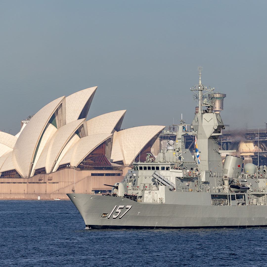 The image shows a naval ship with the number "157" on its bow sailing through the water, with the iconic Sydney Opera House in the background. The ship appears to be a part of the Australian Navy, likely a warship or frigate, with visible radar equipment and military features on its deck. The juxtaposition of the naval vessel and the Sydney Opera House creates a striking contrast between military presence and one of Australia's most recognisable cultural landmarks. The scene takes place on a calm day, with the sky clear and the water serene.