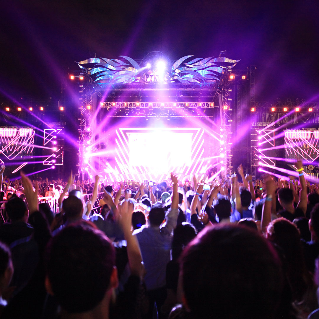 The image shows a vibrant, energetic music festival or concert scene at night. A large crowd of people is gathered in front of a brightly lit stage, with many raising their hands in the air in excitement. The stage is illuminated by purple and pink lights, with intricate light patterns and beams radiating from it. The stage setup includes large screens and decorative elements, adding to the dynamic atmosphere. The audience appears fully engaged, enjoying the live performance in a lively and festive environment. The image captures the essence of a large-scale outdoor music event, filled with excitement and energy.