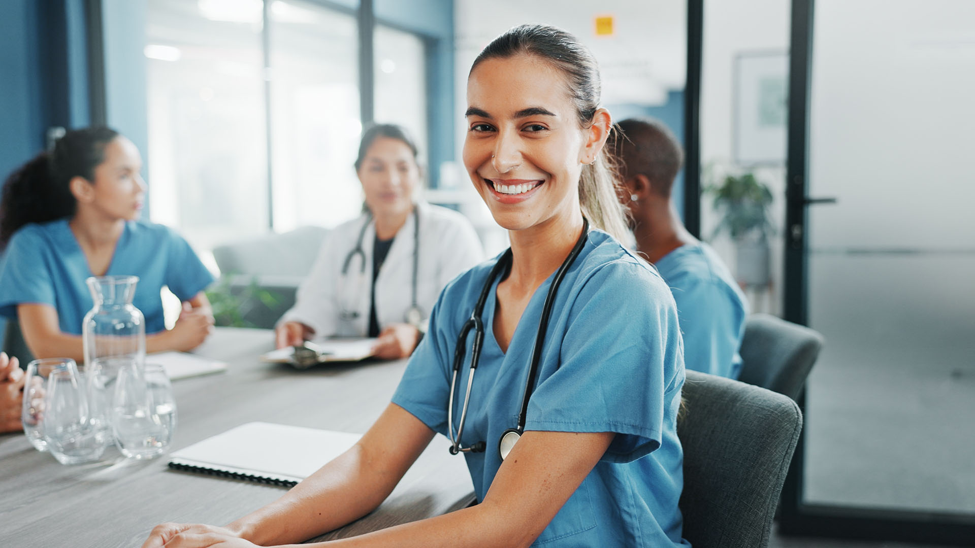 The image shows a group of healthcare professionals in a meeting or discussion, seated around a table. In the foreground, a young woman in medical scrubs with a stethoscope around her neck is smiling at the camera, exuding a positive and friendly demeanor. In the background, other medical staff members, including a doctor and nurses, are engaged in conversation. The setting appears to be a hospital or clinic, with a modern, clean, and professional environment. The image conveys teamwork, collaboration, and a positive working atmosphere in the healthcare field.