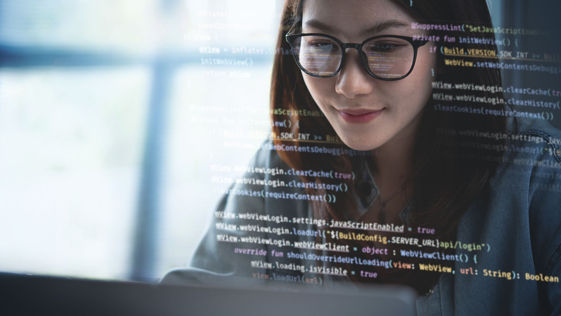 The image shows a young woman with glasses working on a laptop. Her face is focused and illuminated by the screen, with lines of computer code reflecting on her glasses and faintly overlaid on the image. The code appears to be related to web development or programming, hinting that she may be engaged in software or coding tasks. The background is softly blurred, emphasizing her concentration on the work.