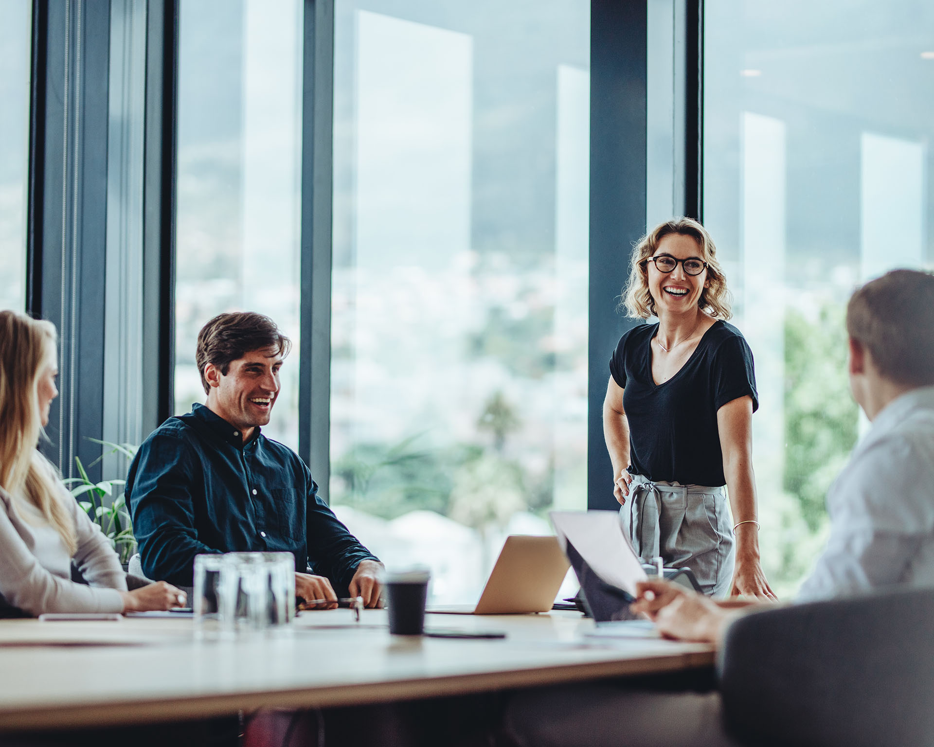 The image shows a group of professionals in a modern office setting, engaged in a casual meeting or discussion. A woman, standing confidently at the head of the table, is smiling and talking to the group, creating an open and collaborative atmosphere. The seated individuals, both male and female, are engaged, listening, and smiling, suggesting a positive and productive environment. Large windows in the background allow natural light to flood the room, offering views of trees and buildings outside. The scene conveys a sense of teamwork, leadership, and a relaxed yet professional work environment.