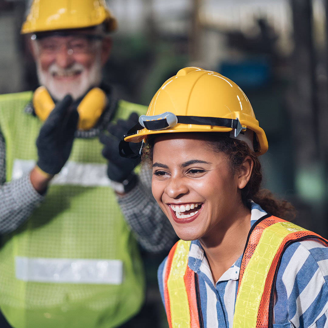 The image shows a young woman wearing a yellow hard hat and safety vest, smiling and laughing in what appears to be a work or industrial environment. Behind her, an older man, also in safety gear with a hard hat, gloves, and a high-visibility vest, is clapping and smiling, suggesting a moment of celebration or recognition. The scene conveys a positive and supportive atmosphere, likely related to teamwork, achievement, or camaraderie in the workplace. The focus is on the joy and encouragement shared among colleagues in a professional, safety-conscious environment.